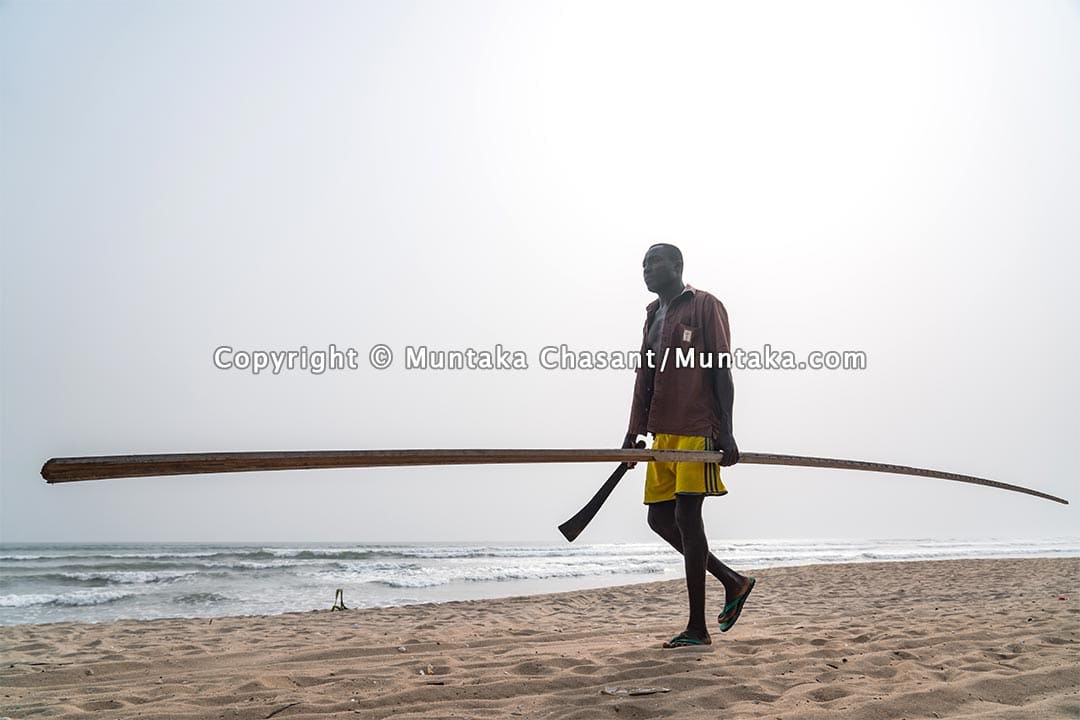 Man walks on the beach holding cutlass and a long stick. Copyright © 2021 Muntaka Chasant