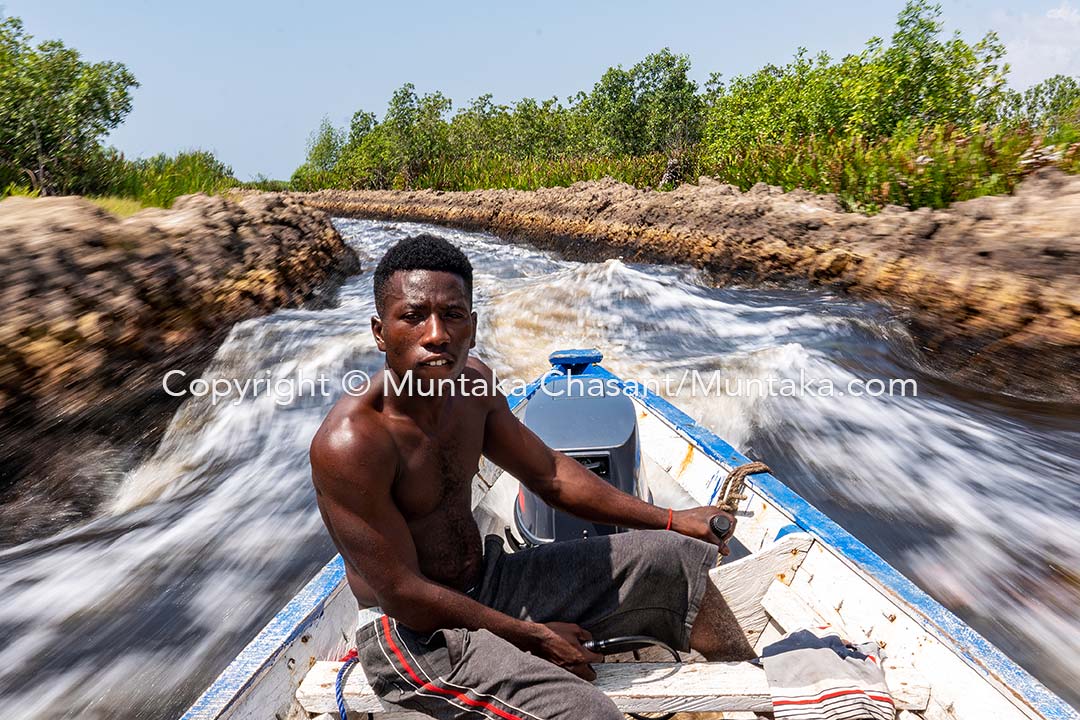 Jacob, a 22-year-old boatman, pilots his motorized canoe against the stream. Copyright © Muntaka Chasant