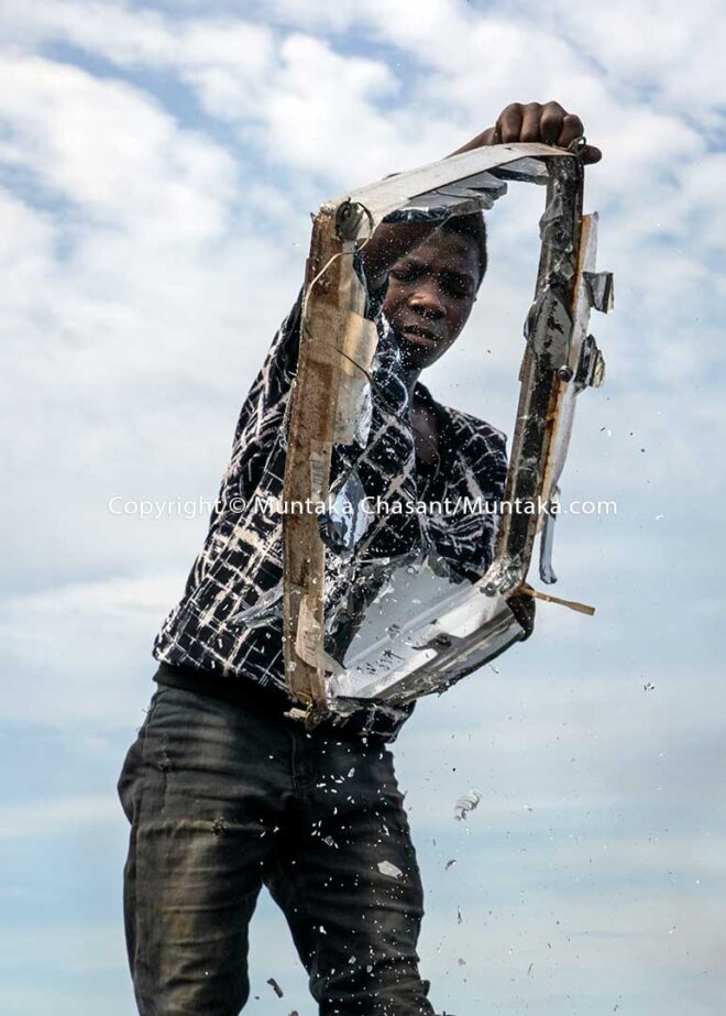E-waste recycling: Osei, 15 years old, shakes off panel glass from an old CRT TV part at Agbogbloshie, Ghana. Old television sets are smashed against rocks sometimes to separate the glasses from the iron metal components inside. They are then sold for around $0.15 per kilo (in May 2020) at Agbogbloshie. © 2020 Muntaka Chasant