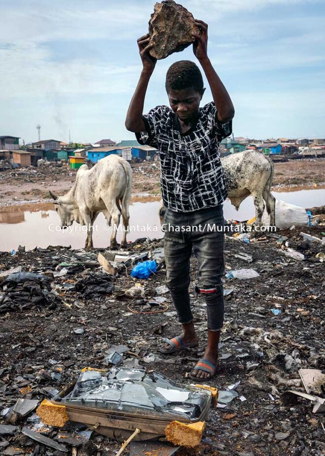 Worst Forms of child labour: 15-year-old Osei is about to smash an old CRT TV against a rock and other CRTs to recover iron materials, Agbogbloshie, Ghana. The metals are sold for around $0.15 per kilo as of May 2020. This worst form of child labour exposes Osei to lead and other toxic substances, including cadmium and barium. The lead in CRT funnel glass is known to pose risks to human health and the environment. © 2020 Muntaka Chasant